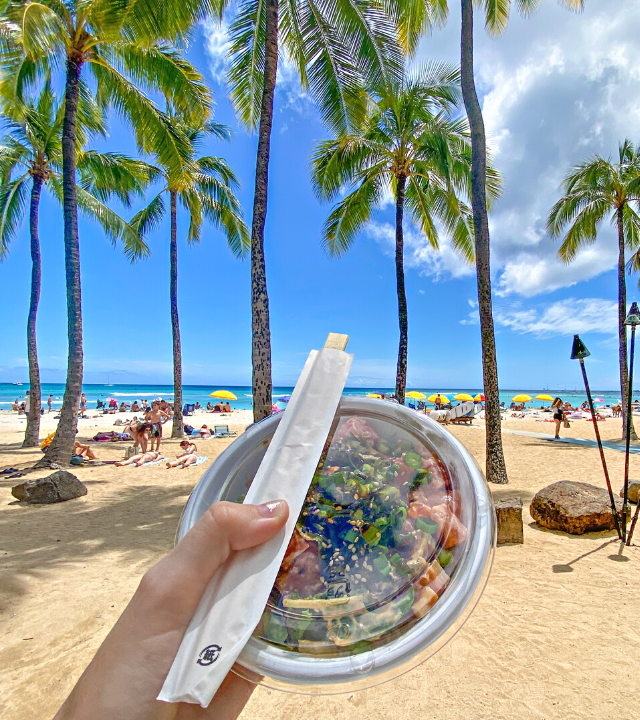Hand holding out poke bowl in front of Waikiki Beach in Oahu, Hawaii