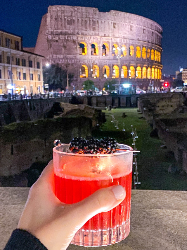 Hand holding a cocktail in front of a view of the Colosseum in Rome at night