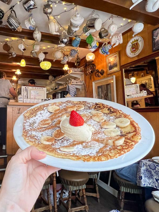 Pancake covered in powdered sugar and bananas on a plate inside Pancakehouse Upstairs in Amsterdam, showing tea pots hanging from the ceiling as decor.