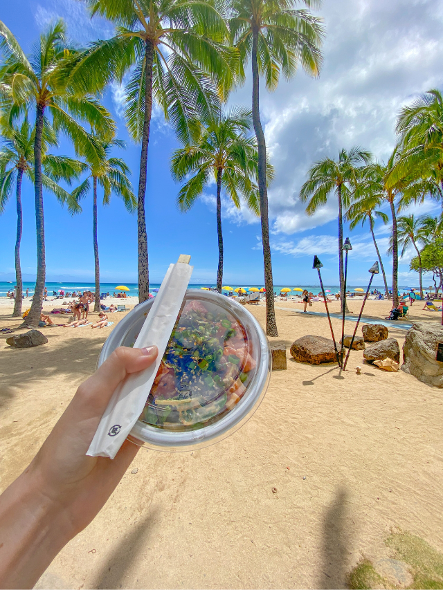 Hand holding out a fresh poke bowl in front of palm trees and sand lining Waikiki Beach in Honolulu, Hawaii